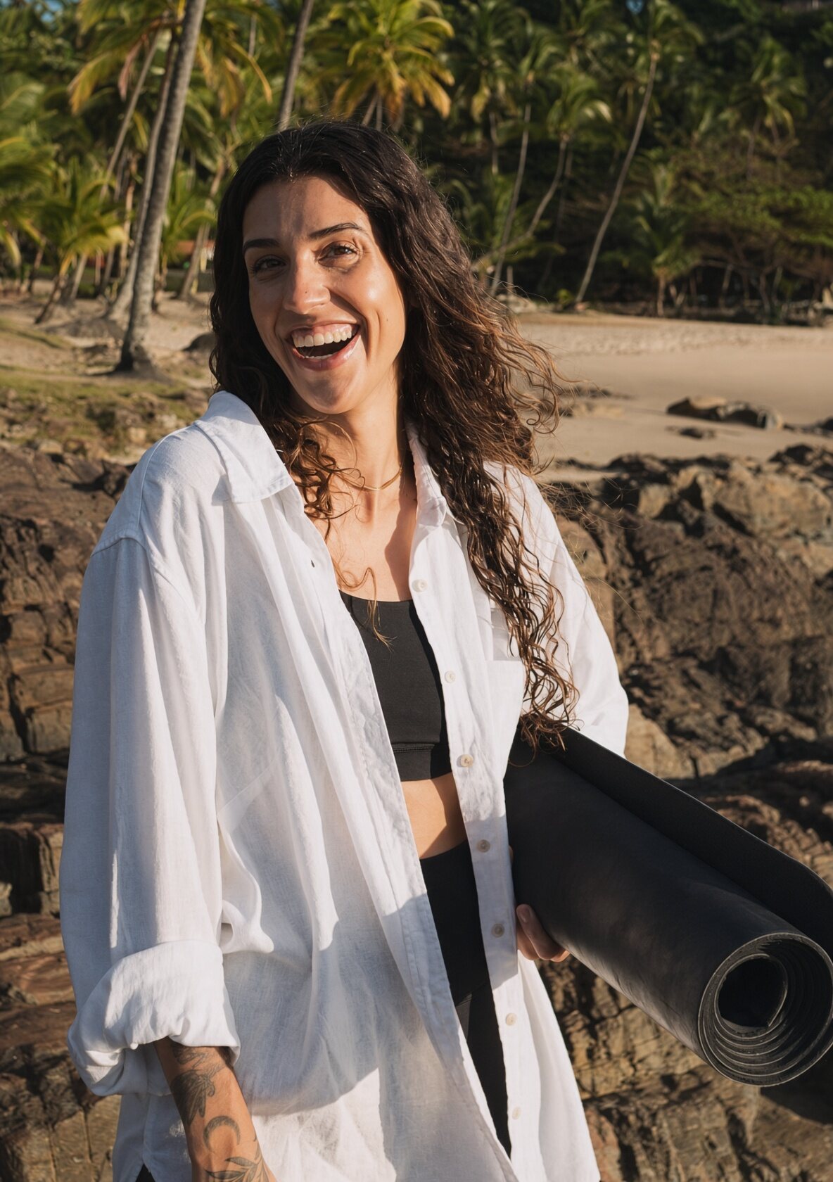 Mayra laughing with her mat under her arm, Praia da Concha behind her